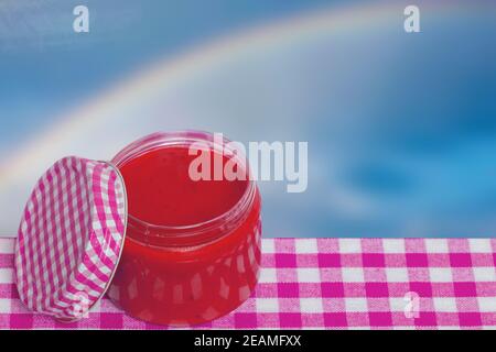 Closeup di un bicchiere aperto di marmellata di fragole fatte in casa su un tavolo su un astratto sfondo arcobaleno. Dima per il montaggio di prodotti alimentari o pubblicità. Foto Stock