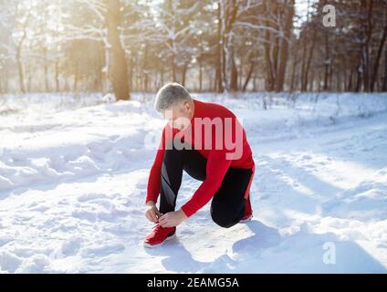 Jogger maturo in caldo sportswear allacciando scarpe su strada innevata al parco invernale Foto Stock