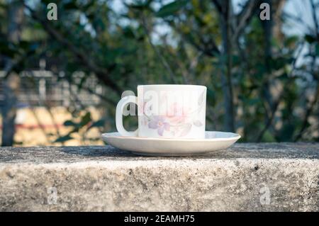 Tazza di caffè alla luce del sole del mattino. Look fresco estivo. Tazza di caffè bianca sul piattino per bevande calde sul tetto di un edificio residenziale con sfondo bokeh. Foto Stock