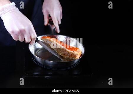 Un pezzo di pesce rosso viene fritto in una padella. Foto su sfondo nero. Persona irriconoscibile. Foto Stock