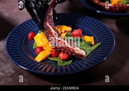 Il cuoco mette un tentacolo di polpo bollito su un piatto. Insalata di polpo con mango e fragole su un piatto blu. Primo piano. Persona irriconoscibile. Foto Stock