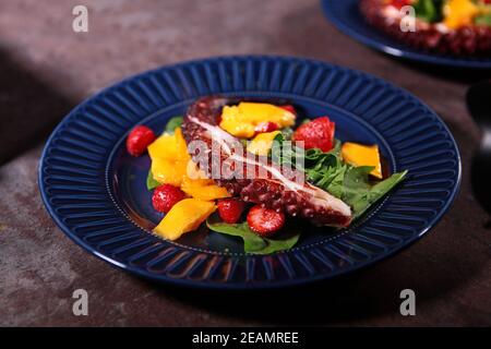 Insalata di polpo con mango e fragole su un piatto blu. Tentacolo di polpo su un piatto blu con frutta e bacche. Primo piano. Foto Stock