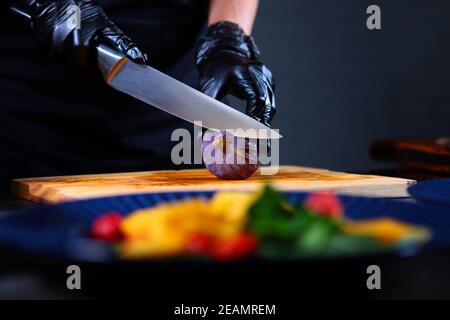 Cuocete in guanti protettivi tagli fichi. Preparazione di ingredienti per insalata di mango e polpo. Persona irriconoscibile. Fuori fuoco. Foto Stock