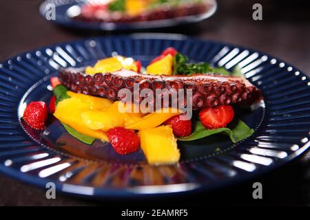 Insalata di polpo con mango e fragole. Tentacolo di polpo su un piatto blu con frutta e bacche. Primo piano. Foto Stock