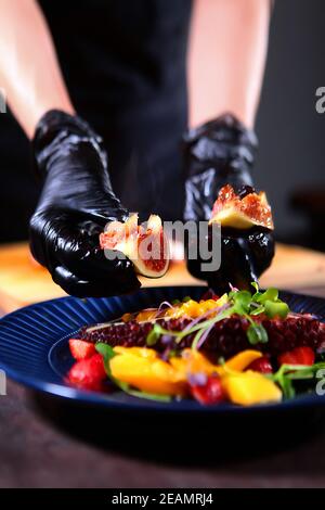 Il cuoco aggiunge fichi all'insalata di polpo e mango. Fuori fuoco. Cucinare gli ingredienti per l'insalata di polpo. Foto verticale. Foto Stock