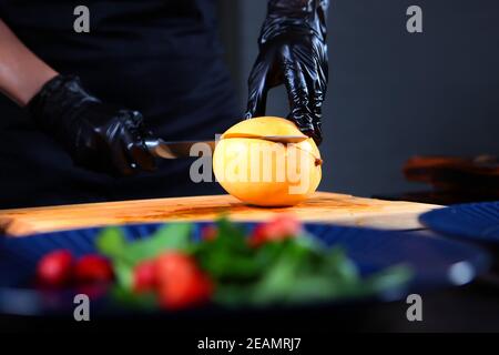 Mango maturo nelle mani dello chef. Cuocete in guanti protettivi tagliano il mango. Preparazione di insalata di mango e polpo. Persona irriconoscibile. Foto all'interno. Foto Stock