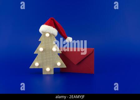 Albero di Natale in legno stilizzato con cappello Babbo Natale in cima  accanto a una busta rossa per i saluti su uno sfondo blu festoso. Capodanno  e Natale saluto Foto stock -