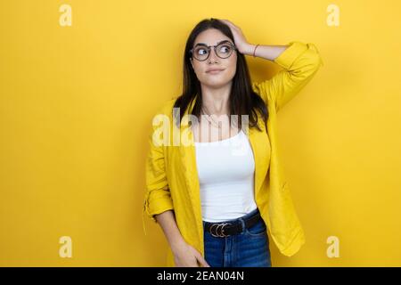 Giovane donna d'affari brunette che indossa blazer giallo su sfondo giallo mettere una mano sulla testa sorridente come aveva dimenticato qualcosa Foto Stock