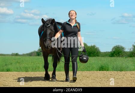 Sorridente donna caucasica, 40 anni, sta tenendo le redini del suo cavallo saddled e camminando sul terreno sabbioso all'aperto. Foto Stock