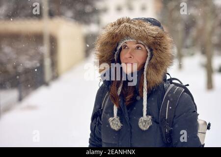 Donna di mezza età con zaino in autunno invernale con pelliccia giacca calda rifinita in piedi in una strada urbana che guarda in su con un sorriso come lei watche Foto Stock