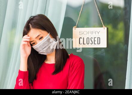 Donna indossare maschera viso proteggere lei triste appendere 'ORRY NOI SONO chiusi, SI PREGA DI TORNARE di nuovo 'bordo alla porta d'ingresso Foto Stock