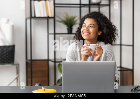 Sorridente giovane gioiosa freelance afroamericana che si geva il caffè del mattino mentre lavorava sul computer portatile, guardando lontano seduto alla scrivania moderna Foto Stock