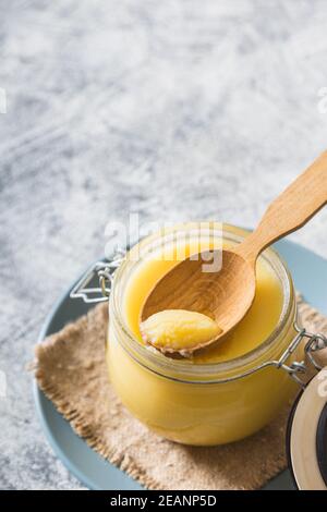 Ghee o burro chiarificato in vaso e cucchiaio di legno su tavola grigia. Vista dall'alto. CopySpace. Il burro di ghee ha grasso sano ed è un ingrediente di cottura comune in molti degli alimenti indiani Foto Stock