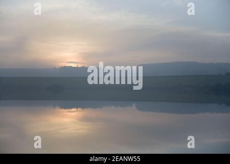 Tramonto color pastello dietro gli alberi sopra l'acqua del bacino idrico. Nuvole frizzanti Foto Stock