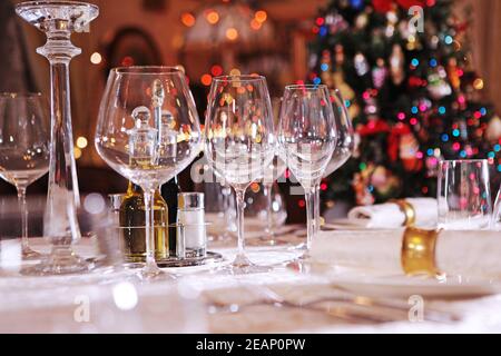 Impostazione del tavolo per le vacanze Capodanno o Natale. Albero di Natale fuori fuoco. Bicchieri di vino in vetro vuoti. Foto Stock