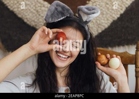 Buona Pasqua! Felice giovane donna in spighe conigliate e vestito di lino che tiene le uova di pasqua agli occhi e sorridente sullo sfondo della stanza rustica. Caste tinte naturali Foto Stock