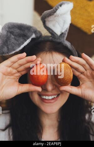 Buona Pasqua! Felice giovane donna in spighe conigliate e vestito di lino che tiene le uova di pasqua agli occhi e sorridente sullo sfondo della stanza rustica. Caste tinte naturali Foto Stock