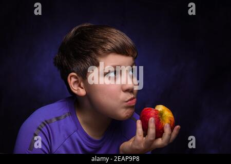 ragazzo bianco con capelli marrone chiaro, vestito con una t-shirt sportiva viola, mastica e tiene in mano una grande mela rossa e gialla su uno sfondo isolato Foto Stock