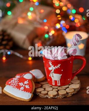 Coppa in ceramica rossa con cacao e marshmallows e pan di zenzero al forno, luci di Natale dietro Foto Stock