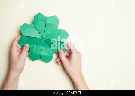 Trifoglio a quattro foglie di Origami, in carta verde, tenuto da due mani di giovane donna su uno sfondo chiaro. Concetti di fortuna e giorno di San Patrizio. Immagine w Foto Stock