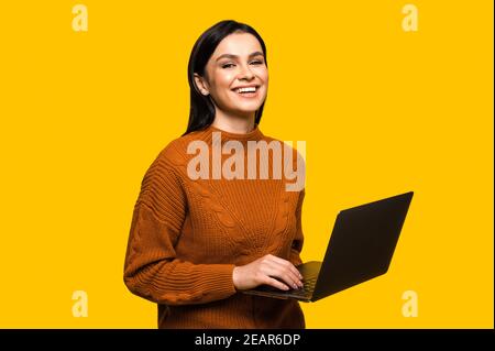 Soddisfatto attraente giovane caucasica sorridente donna in felpa casual, si leva su isolato sfondo arancione, tiene e usa, guarda e sorride alla macchina fotografica Foto Stock