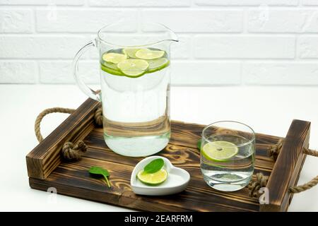 Una limonata fatta in casa fatta di lime si trova in un bicchiere e caraffa su un vassoio di legno Foto Stock