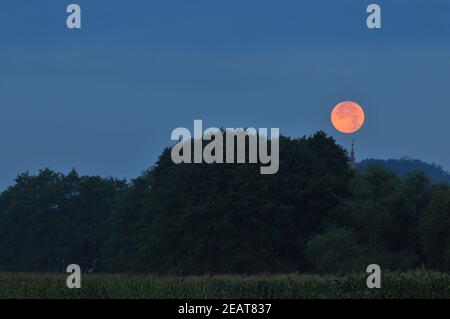 Luna piena che sorge sopra il campanile della chiesa durante il tramonto estivo Foto Stock