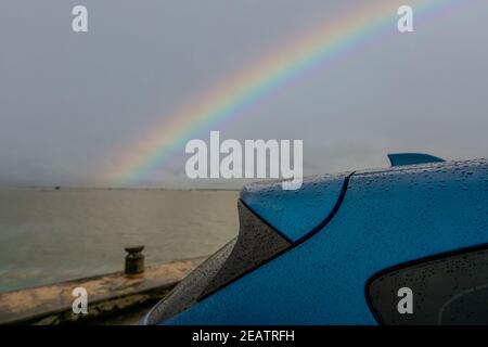 Fuoco selettivo sul tronco dell'automobile blu di lusso del SUV con le gocce di pioggia sull'arcobaleno e sullo sfondo del cielo tempestoso. Auto parcheggiata nel parcheggio esterno accanto alla spiaggia di mare in giornata piovosa. Foto Stock