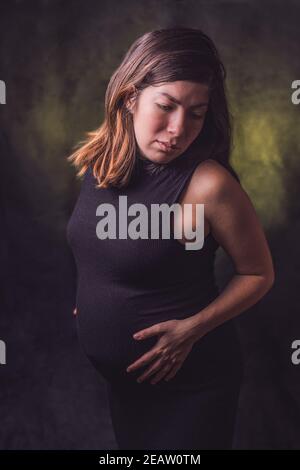 Ritratto di una donna incinta dai capelli scuri in una stretta nera vestito tenendo la sua grande ventre gestazione con le mani Foto Stock