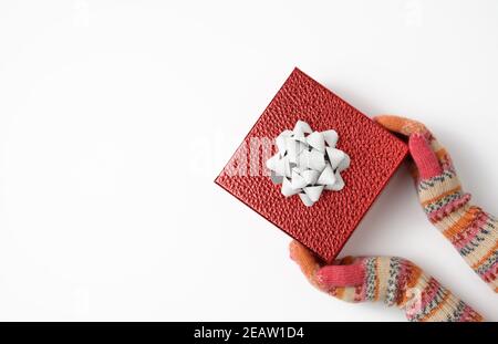 le mani femminili in guanti lavorati a maglia tengono una scatola rossa sopra uno sfondo bianco Foto Stock