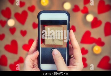 Telefono cellulare femminile che tiene la mano scattando la foto della scatola del regalo per San Valentino, sfondo sfocato, biglietto d'auguri romantico presente di fondo Foto Stock