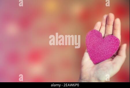 Mano femminile che tiene il cuore rosa glitter su sfondo romantico bokeh sfocato, spazio di copia o spazio per il testo, San Valentino o sfondo romantico Foto Stock