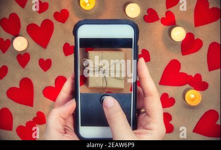Telefono cellulare femminile che tiene la mano scattando la foto della scatola del regalo per San Valentino, sfondo sfocato, biglietto d'auguri romantico presente di fondo Foto Stock
