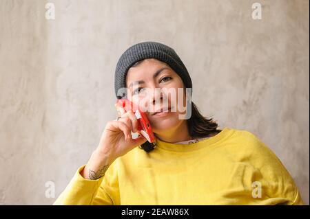 una giovane bruna sorridente carina con un cappuccio a maglia e una felpa gialla che parla su un cellulare in un caso rosso Foto Stock