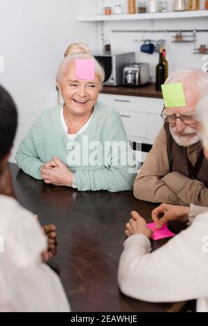 amici anziani felici e multiculturali con note appiccicose sulla fronte gioco in cucina Foto Stock