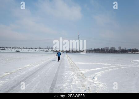 Vista soleggiata all'aperto, un uomo fa sentiero che corre su uno strato spesso neve coperta strada, terra e campo in campagna in Germania durante la stagione invernale. Foto Stock