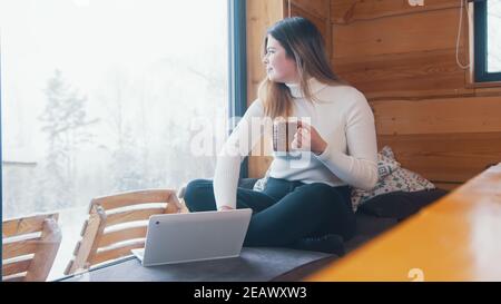 Giovane donna caucasica che beve caffè e che ha videochiamata vicino alla finestra. Girare il laptop per mostrare la neve. Foto di alta qualità Foto Stock