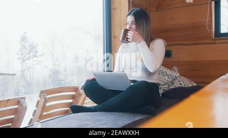 Giovane donna caucasica che beve caffè e usa un computer portatile vicino alla finestra. Vacanze invernali. Foto di alta qualità Foto Stock