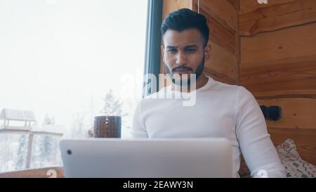 Giovane indiano che beve caffè e lavora su un computer portatile vicino alla grande finestra. Foto di alta qualità Foto Stock