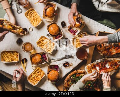 Cena fast food per famiglie con Lockdown dal servizio di consegna a domicilio. Piatto-Lay di gruppo di amici mangiare hamburger, patatine fritte, panini, pizza, bere birra a casa festa sullo sfondo del tavolo, vista dall'alto Foto Stock