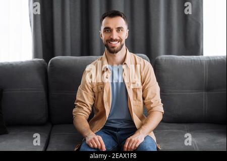 Ritratto di un attraente ragazzo caucasico vestito in elegante abbigliamento casual. Uomo sicuro guarda direttamente la fotocamera con un sorriso amichevole mentre si siede Foto Stock