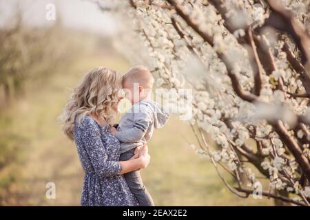 Primo piano ritratto di giovane madre con figlio biondo che si agguantano, donna bacia ragazzo con amore materno tra i giardini fiorenti di fiori bianchi Foto Stock