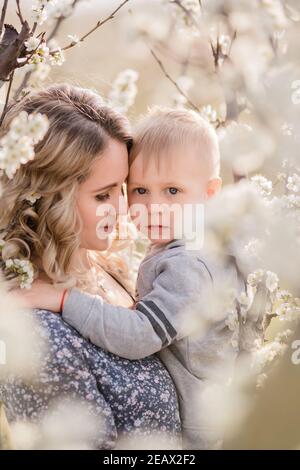 Primo piano ritratto di giovane madre con figlio biondo che si agguantano, donna bacia ragazzo con amore materno tra i giardini fiorenti di fiori bianchi Foto Stock