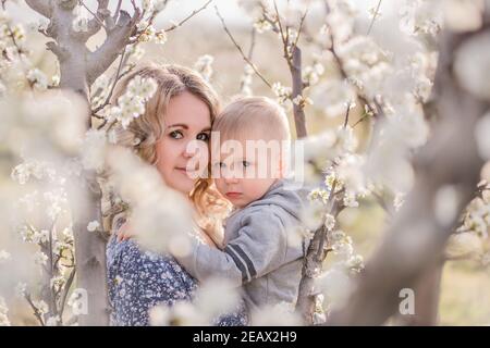 Primo piano ritratto di giovane madre con figlio biondo che si agguantano, donna bacia ragazzo con amore materno tra i giardini fiorenti di fiori bianchi Foto Stock