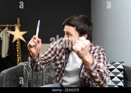 Un giovane ragazzo che balla con gioia mentre si guarda al test di gravidanza. L'uomo non pronto diventa padre e felice di vedere test di gravidanza negativo. Maschio eccitato e. Foto Stock