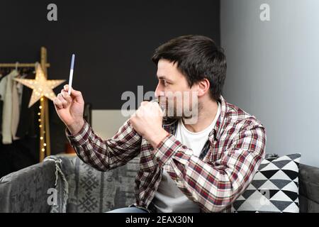 Giovane maschio caucasico felice guardando al test di gravidanza molto felice. Bell'uomo sorridente ispirato e felice e danze, canta mentre si siede sul divano Foto Stock