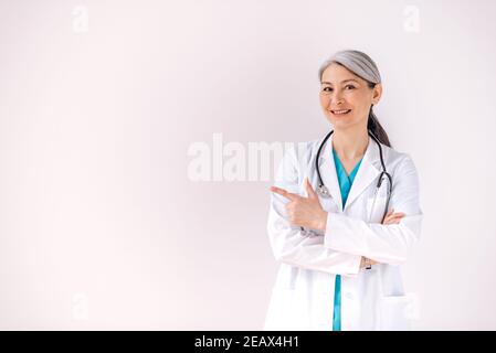 Ritratto di un medico di media età con capelli grigi asiatici in uniforme medica e stetoscopio si trova su sfondo bianco isolato, punta il dito Foto Stock