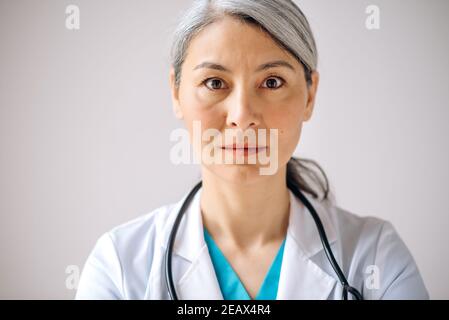 Ritratto in primo piano di una dottoressa asiatica con capelli grigi, medico generico o pediatra in uniforme medica e stetoscopio, in piedi contro l'isolat Foto Stock