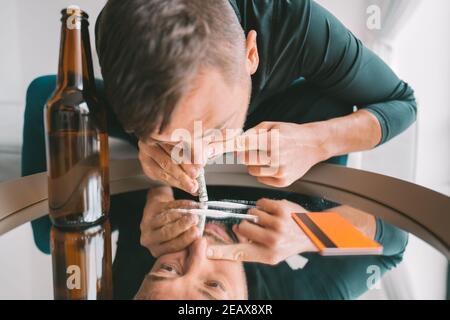 Giovane dipendente junkie uomo sniffing o russare linee di cocaina su specchio attraverso banconote arrotolate. Narcotici e concetto di dipendenza. Foto Stock