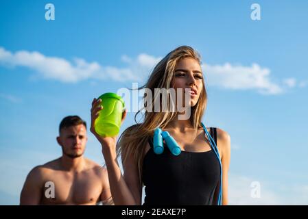 Slim allenatore di donna fitness con shaker proteine. Sorridente giovane donna e allenatore di coppia personale. Uomo e ragazza con bottiglia di frullato proteico e saltellando Foto Stock